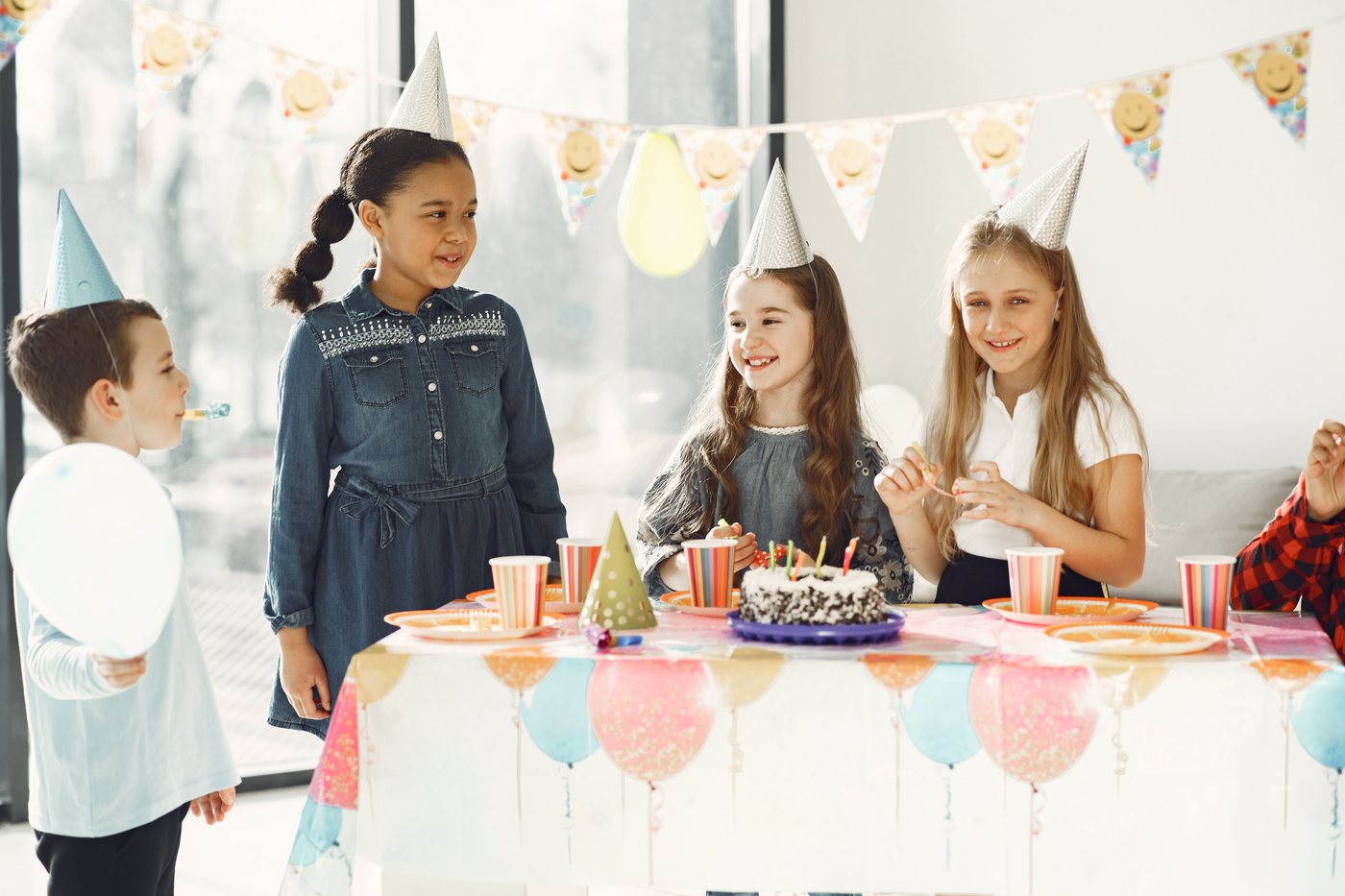 Children in party hats gathered around a birthday table with cake and balloons