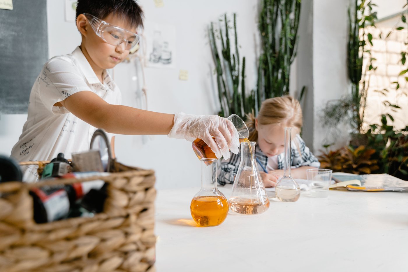A boy in safety glasses pours liquid into a flask while a classmate works beside him